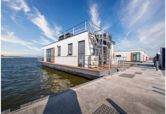 Modern houseboat in Ribnitz-Damgarten with rooftop terrace, floating on water alongside a dock in sunlight.