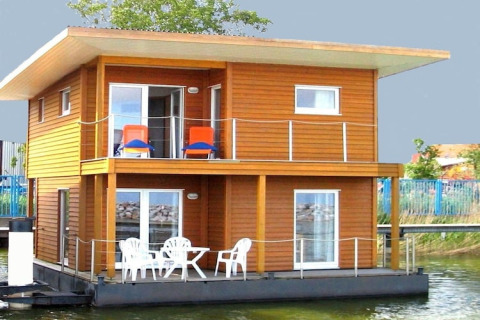 Modernes Hausboot mit Holzverkleidung, zwei Etagen, Balkon und Gartenmöbeln, am Ufer vertäut.