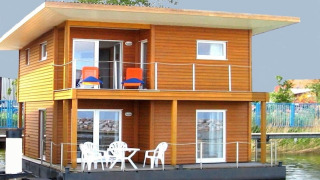 Modernes Hausboot mit Holzverkleidung, zwei Etagen, Balkon und Gartenmöbeln, am Ufer vertäut.