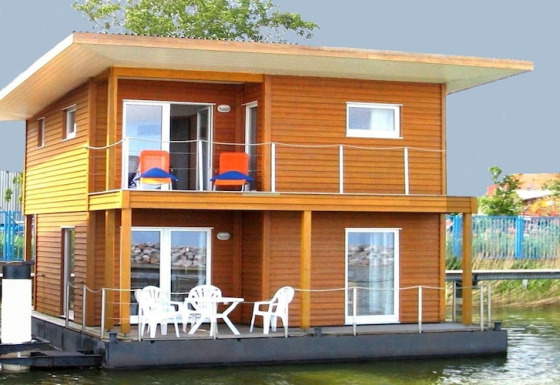 Modernes Hausboot mit Holzverkleidung, zwei Etagen, Balkon und Gartenmöbeln, am Ufer vertäut.