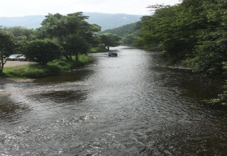 A vehicle drives through a shallow river near a glamping site, surrounded by lush trees and distant hills.