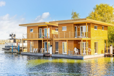 Maison flottante moderne en bois avec balcons et chaises blanches, amarrée dans un port ensoleillé.