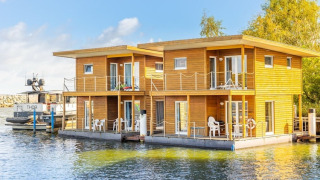 Modernes Hausboot mit Holzverkleidung und Balkonen, festgemacht an einem sonnigen Hafentag.