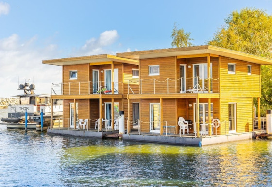 Moderne woonboot met houten gevel, balkons en witte stoelen, aangemeerd in een zonnige haven.