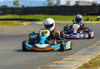 To gokartkørere på en bane med hjelme og dragter, en førende og en anden tæt bagefter i et løb.