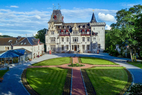 Et smukt slotlignende lodge, La Ribambelle, i Belgien, omgivet af velplejede haver under en blå himmel.
