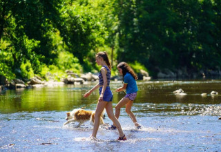 To piger og en hund leger i en lav flod omgivet af grøn natur ved La Ribambelle-lodgen i Belgien.