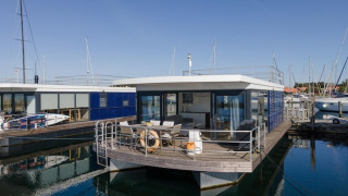 Foto af en moderne husbåd med stor terrasse, glasfacader og udsigt til havnen på en solskinsdag.