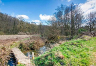 Træbro over en bæk i naturligt landskab ved La Ribambelle-lodgen i Belgien under blå himmel.