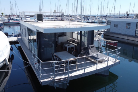 Moderne husbåd med stor terrasse og glasvægge ligger fortøjet i marina omgivet af sejlbåde.