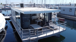 Moderne husbåd med stor terrasse og glasvægge ligger fortøjet i marina omgivet af sejlbåde.