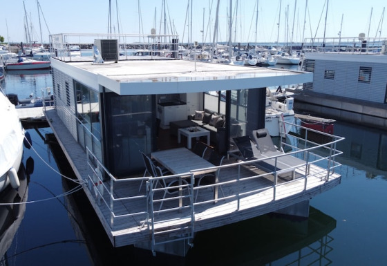 Moderne husbåd med stor terrasse og glasvægge ligger fortøjet i marina omgivet af sejlbåde.