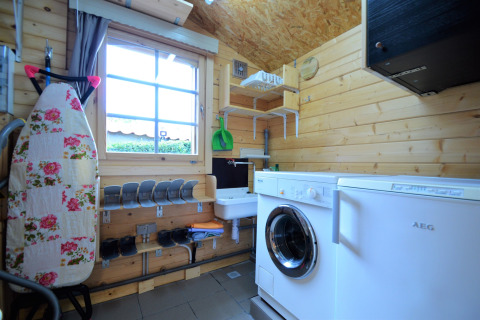 Buanderie dans un chalet en bois avec lave-linge, table à repasser fleurie et grande fenêtre en Belgique.