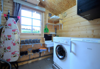Buanderie dans un chalet en bois avec lave-linge, table à repasser fleurie et grande fenêtre en Belgique.