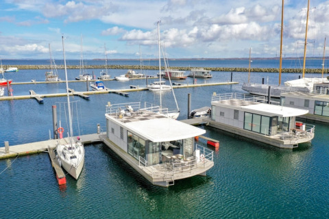 Houseboat moderne e barche a vela sono ormeggiate in un porto turistico sotto un cielo blu nuvoloso.