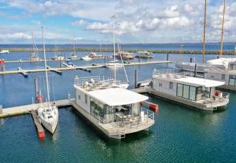 Des péniches modernes et des voiliers sont amarrés dans une marina sous un ciel bleu partiellement nuageux.
