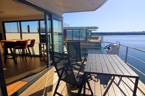 Modern houseboat with a deck, chairs, and table overlooking calm water and nearby houseboats.