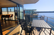 Udsigt fra en husbåd med terrasse, stole og bord mod roligt vand og andre husbåde i baggrunden.
