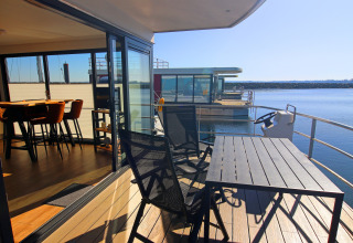 Modern houseboat with a deck, chairs, and table overlooking calm water and nearby houseboats.