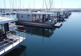 Moderni houseboat ormeggiati in un porto calmo, circondati da barche a vela e acqua limpida e riflettente.