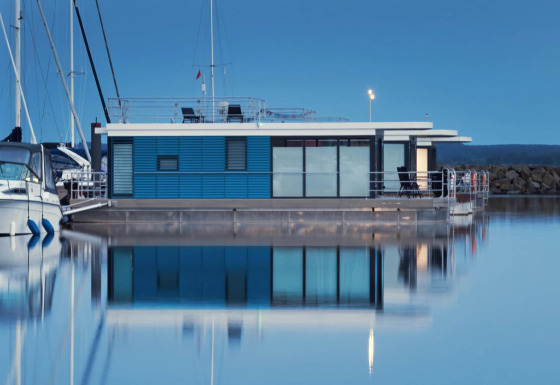 Modern houseboat Family & Friends mit Hund LP 10 moored in a calm marina, reflected in still blue water.