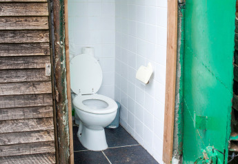Udendørs toilet på en husbåd med åbne døre, hvide fliser, trævæg og grøn metalramme i Belgien.