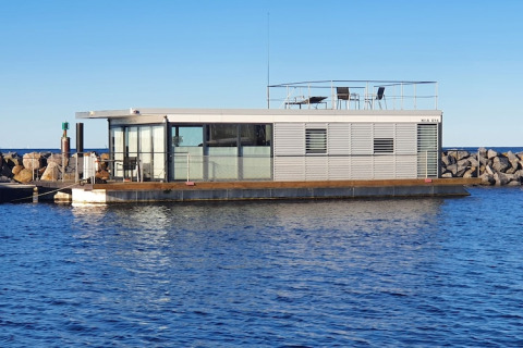 Péniche moderne amarrée près d’une jetée rocheuse, avec grandes vitres et terrasse sur le toit.