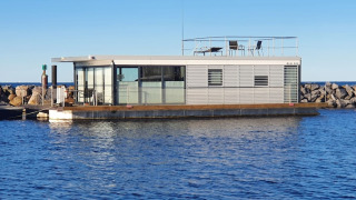 Péniche moderne amarrée près d’une jetée rocheuse, avec grandes vitres et terrasse sur le toit.