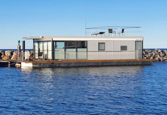 Péniche moderne amarrée près d’une jetée rocheuse, avec grandes vitres et terrasse sur le toit.