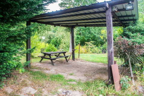 Overdekte picknickplek met houten tafel en bank in een groene tuin bij een woonboot in België.