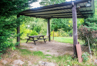 Overdekte picknickplek met houten tafel en bank in een groene tuin bij een woonboot in België.