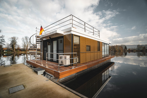 Modern houseboat named Houseboat in Höxter docked at the pier, with a German flag and calm water.