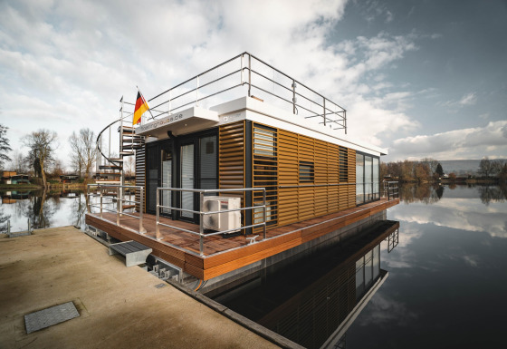 Casa flotante moderna en Höxter, atracada en el muelle con bandera alemana y agua tranquila alrededor.