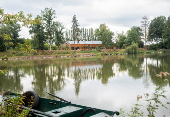 Casa galleggiante sul lago privato in Belgio, con barca verde in primo piano e alberi riflessi sull’acqua.