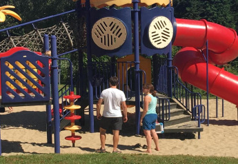 Two adults stand by a colorful playground with slide at Holiday Park Ardinam 1 in Belgium on a sunny day.