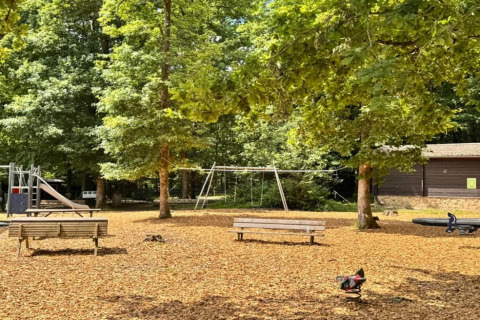 Parque infantil junto al chalet 'OREE DU BOIS' en Bélgica, con columpios, tobogán y bancos rodeados de árboles.