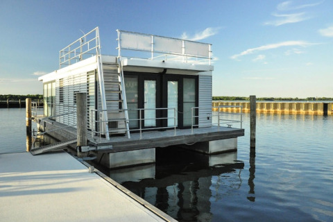 Moderne husbåd kaldet Floating House 'Leni' ligger fortøjet ved kaj i Ribnitz-Damgarten under blå himmel.
