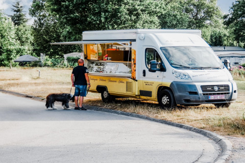 Uomo con cane vicino a un food truck giallo al Holiday Park Ardinam 1, Belgio, circondati dal verde.