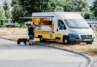 Uomo con cane vicino a un food truck giallo al Holiday Park Ardinam 1, Belgio, circondati dal verde.
