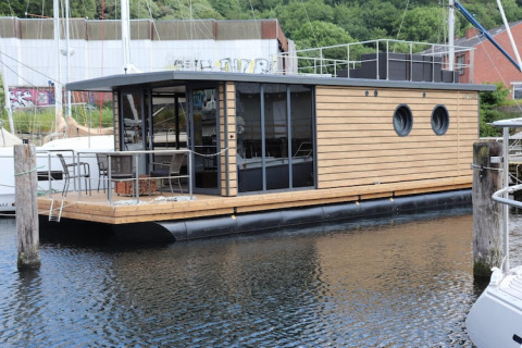 A modern houseboat named Houseboat Leni docked at a marina, featuring wood paneling and outdoor seating.