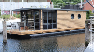 A modern houseboat named Houseboat Leni docked at a marina, featuring wood paneling and outdoor seating.
