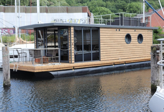 Een eigentijdse woonboot, Houseboat Leni, ligt aangemeerd aan de steiger met houten terras en stoelen.