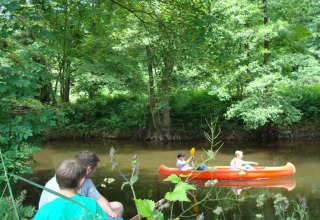 Twee mensen zitten aan de oever en kijken naar twee anderen in een oranje kano op een bosrijke rivier.