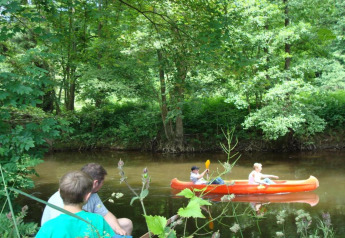 Two people sit on a riverbank while watching two others paddle an orange canoe through a forested area.
