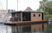 Casa flotante moderna llamada Houseboat Lina con paneles de madera, atracada en un puerto de Alemania.