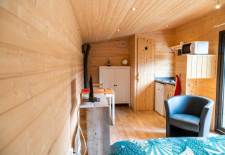 Interno della casa mobile 'Holiday Park Ardinam 1' in Belgio con pareti in legno, angolo cottura e poltrona.