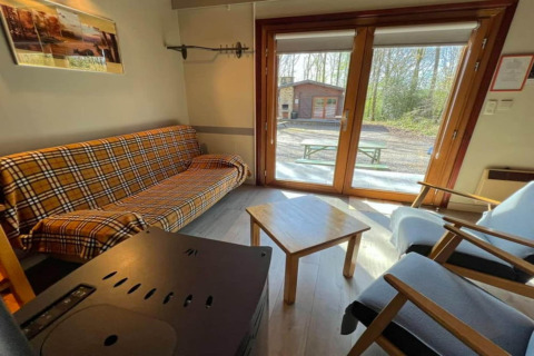 Intérieur du chalet LIJSTERNEST en Belgique, avec canapé à carreaux, table en bois et vue sur la forêt.