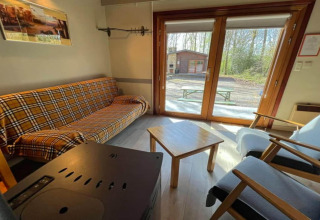 Intérieur du chalet LIJSTERNEST en Belgique, avec canapé à carreaux, table en bois et vue sur la forêt.