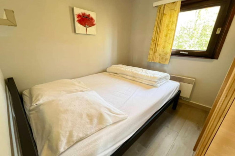 Petite chambre avec un lit double, draps blancs, une fenêtre et un tableau de fleur rouge au mur.