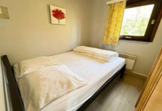 Petite chambre avec un lit double, draps blancs, une fenêtre et un tableau de fleur rouge au mur.